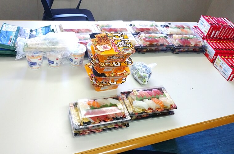 カップラーメンやお寿司、お菓子が机に並べられている写真